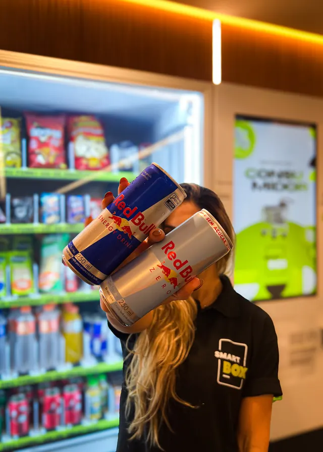 Atendente SmartBox segurando latas de Red Bull em frente à geladeira